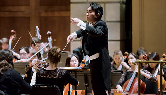 an asian man is standing in the middle of a hall as he is leading an orchestra of students