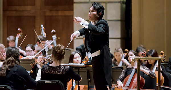 an asian man is standing in the middle of a hall as he is leading an orchestra of students