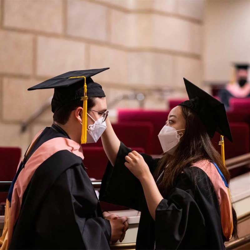 woman helping man with cap/gown during graduation wearing masks in eastman theatre