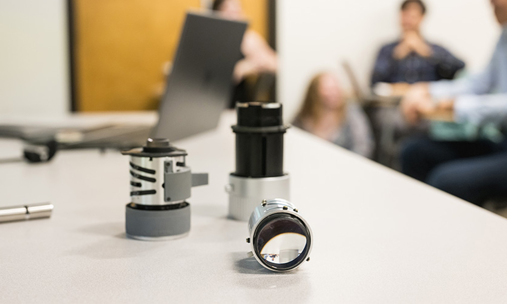Photo of lenses on a white desk with a laptop and students in background