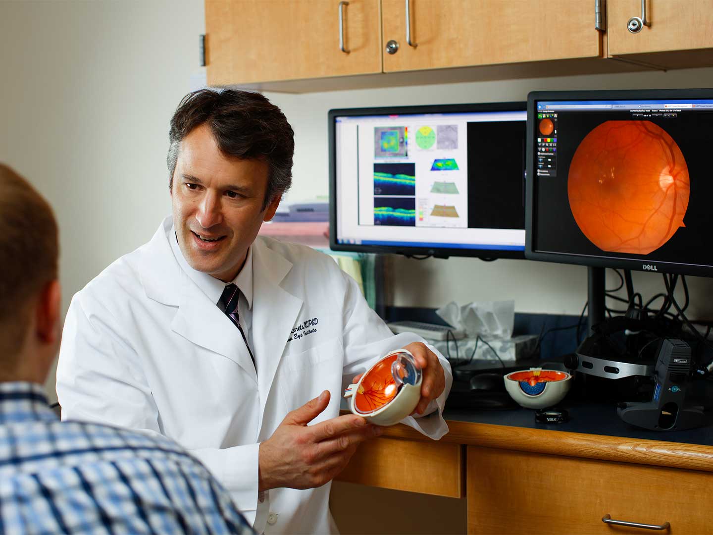 doctor speaking to patient with a model of the eye in his hands and computers in the background with imaging of the internals of an eye