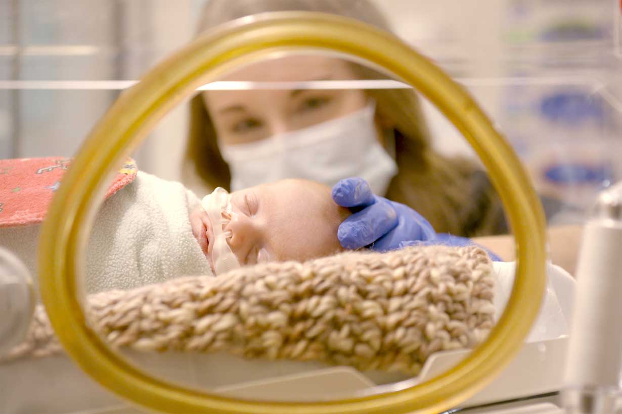 a mom wearing a mask is seen reaching through an incubator with a gloved hand to touch her sleeping baby who is swaddled and has a tube taped to their face