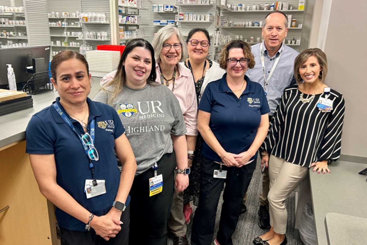 Group of pharmacists pose for a picture, standing in front of the counter, with shelves of medications behind them