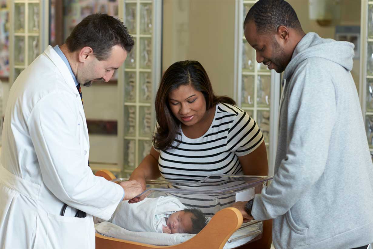 a mother, father, and doctor look down at a swaddled newborn baby in a clear hospital bassinet