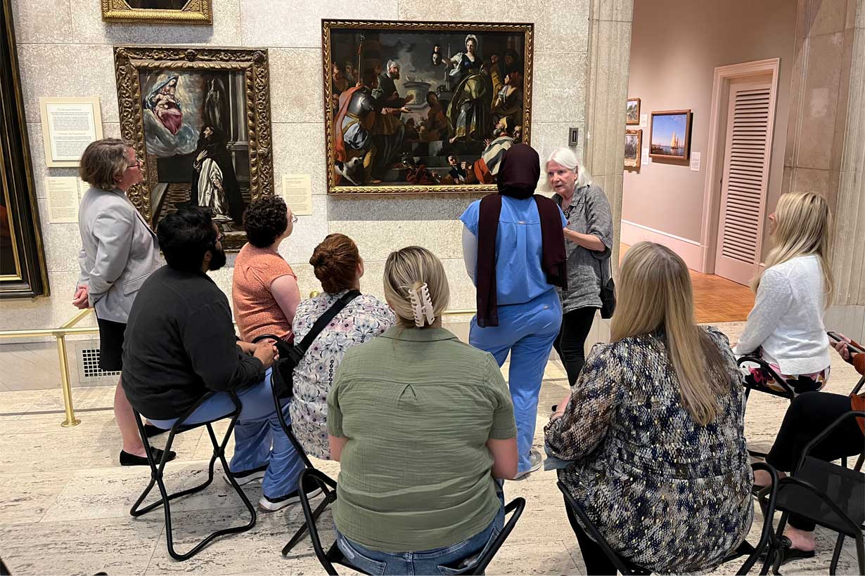 Guide stands in front of framed art works, talking to many seated people, one standing