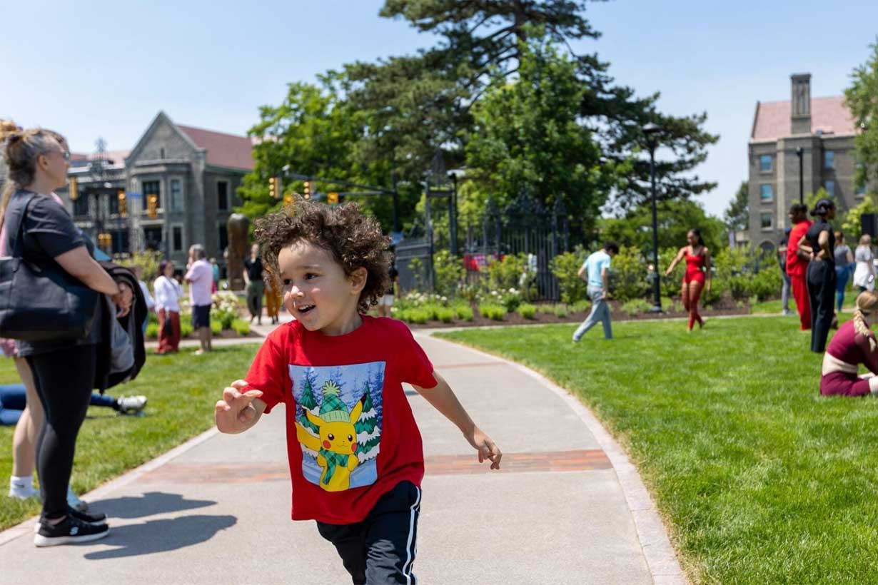 child runs along sidewalk in sculpture garden, several people standing and sitting in the grass