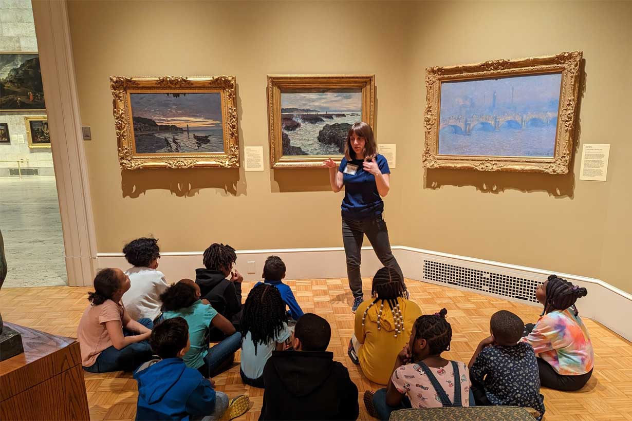 Guide stands in front of three framed art works, talking to many kids seated on the floor