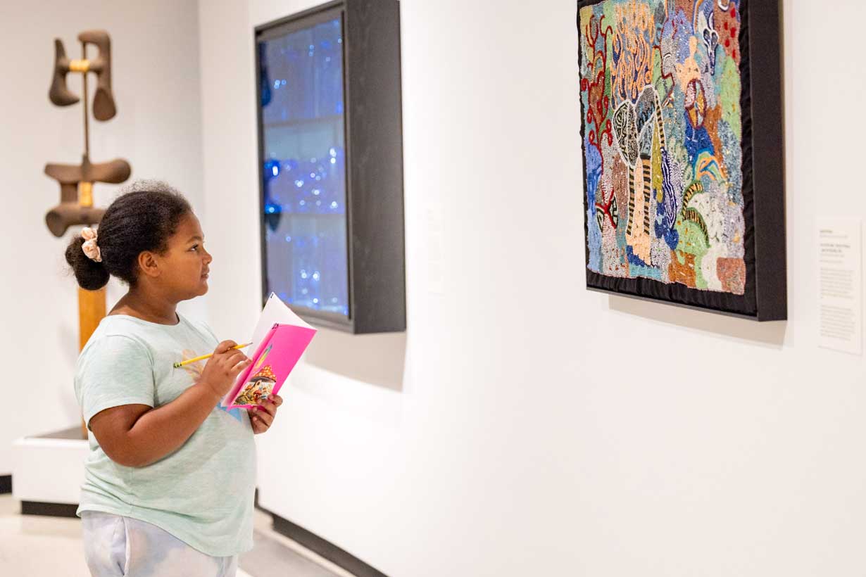 young girl looks at hung artwork, holding an open notebook and pencil