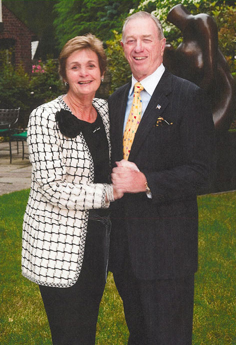 A smiling couple, Bob and Pam Goergen, stand together holding hands on a garden lawn with lush greenery and a sculpture in the background.