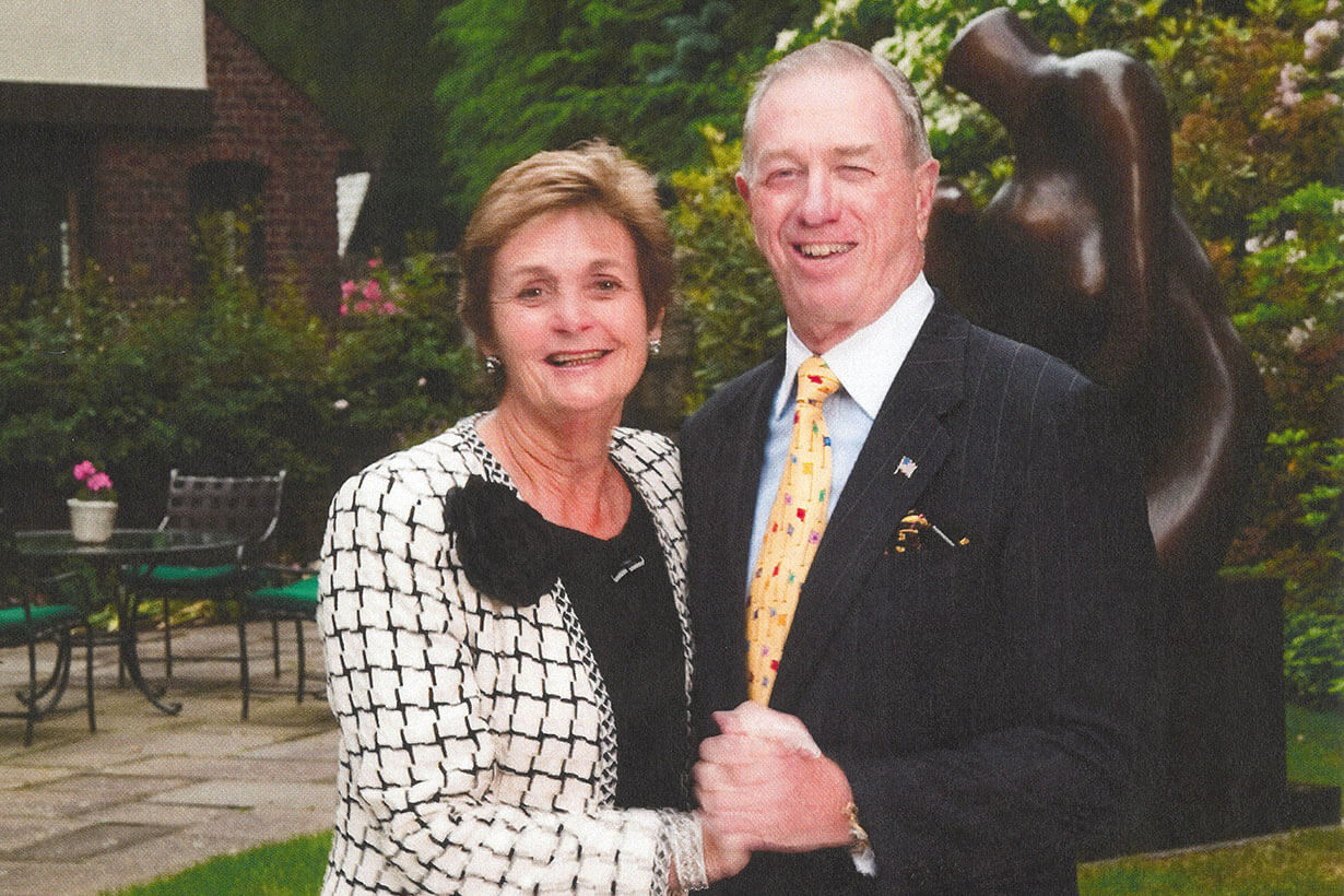 A smiling couple, Bob and Pam Goergen, stand together holding hands on a garden lawn with lush greenery and a sculpture in the background.