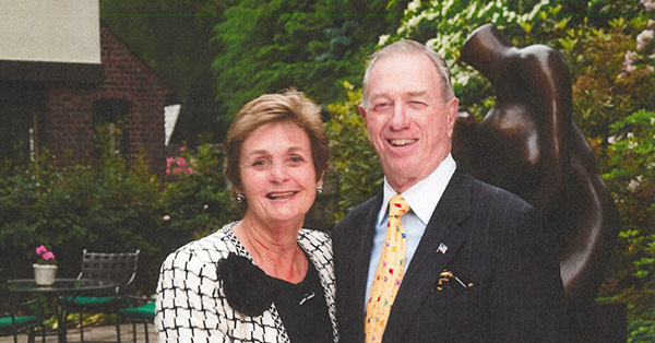 A smiling couple, Bob and Pam Goergen, stand together holding hands on a garden lawn with lush greenery and a sculpture in the background.