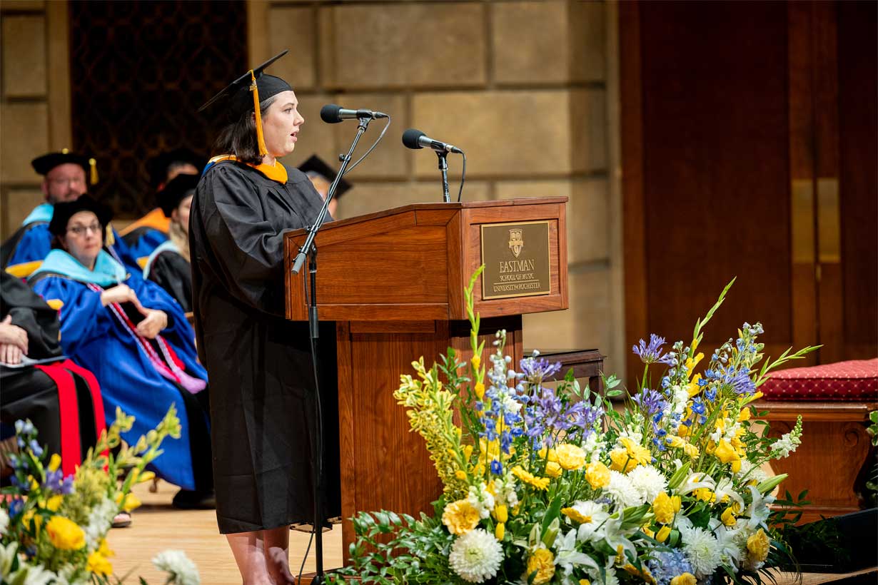 Sarah Mullen stands at a podium in a cap and gown, giving a speech