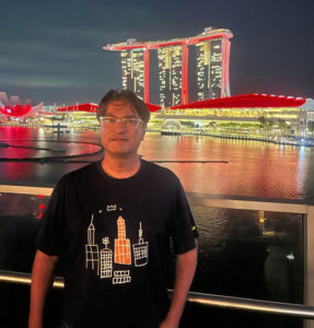 Omar Asghar ’94 standing on a waterfront balcony at night with the illuminated Marina Bay Sands hotel and surrounding Singapore skyline in the background.