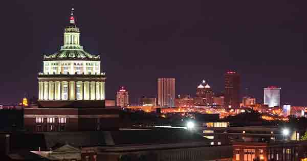 Rush Rhees library tower lighted up at night with Rochester skyline in background