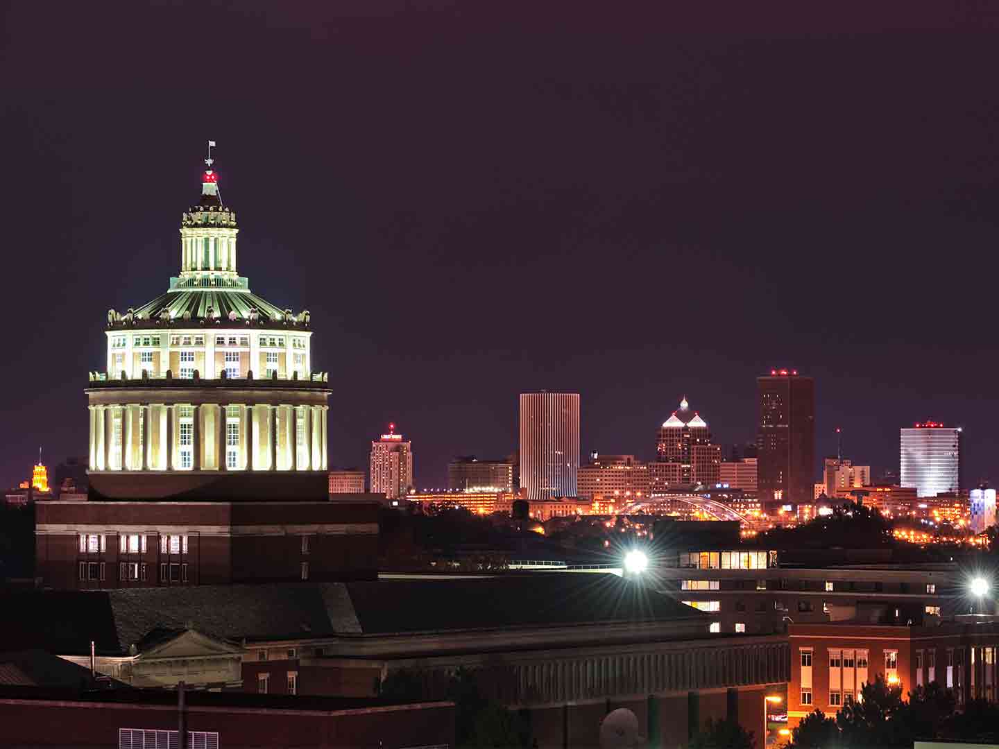 Rush Rhees library tower lighted up at night with Rochester skyline in background
