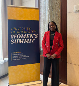 Victoria Liverpool stands beside a University of Rochester Women’s Summit sign, wearing a red blazer and black outfit at the event.