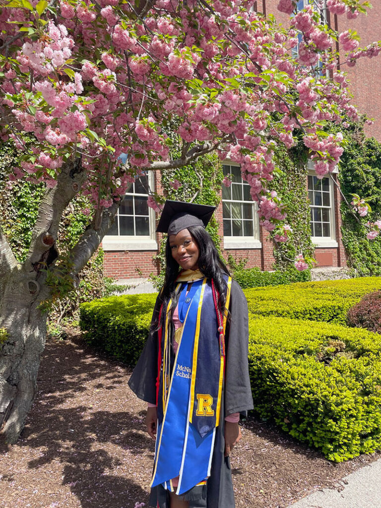 Victoria Liverpool stands smiling beneath a blooming pink cherry blossom tree in front of a campus building during her 2022 graduation from the University of Rochester.
