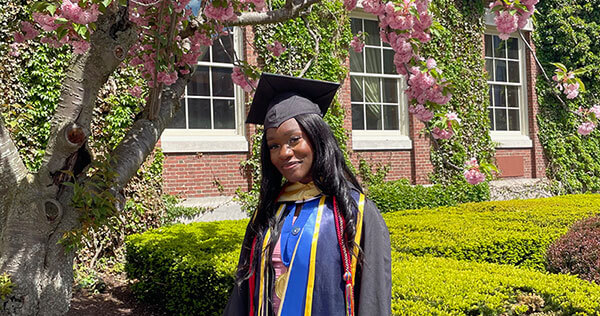 Victoria Liverpool stands smiling beneath a blooming pink cherry blossom tree in front of a campus building during her 2022 graduation from the University of Rochester.
