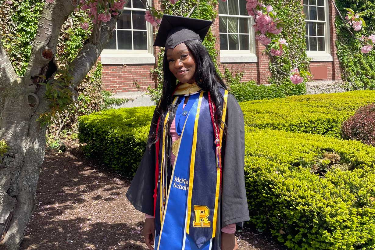 Victoria Liverpool stands smiling beneath a blooming pink cherry blossom tree in front of a campus building during her 2022 graduation from the University of Rochester.
