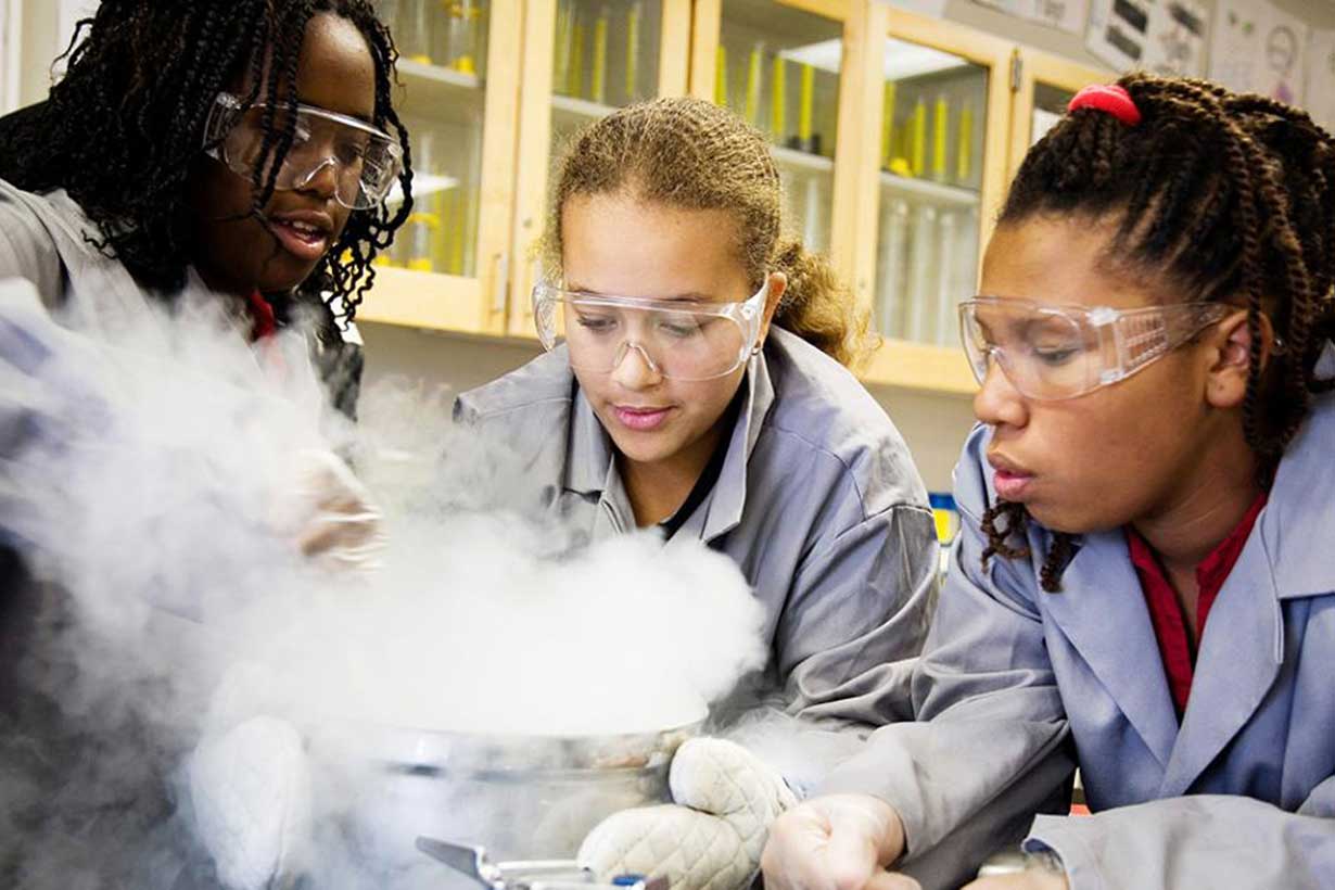 three female students wearing lab coats and goggles look at metal bowl with smoke billowing out