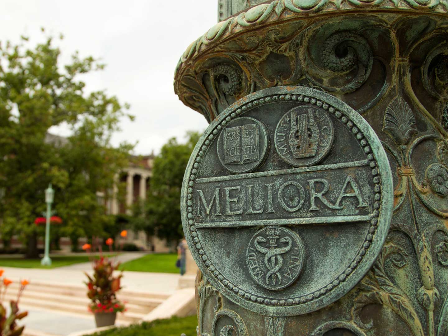 meliora seal at the base of the flag pole, trees, pillared building, and stairs in the background
