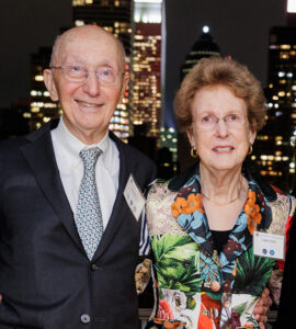 Arnold and Anne Lisio smiling at the camera, with a nighttime city skyline illuminated behind them.