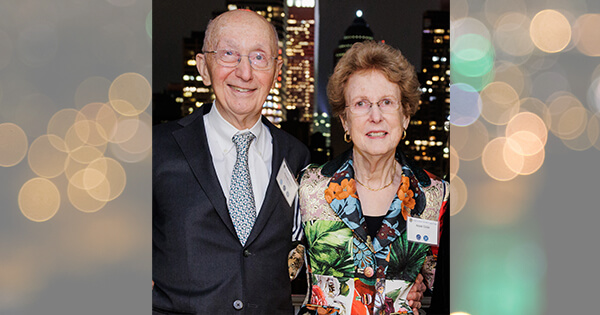 Arnold and Anne Lisio smiling at the camera, with a nighttime city skyline illuminated behind them.
