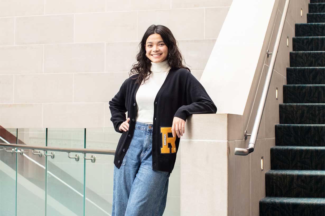 Ashni Budge, standing, leaning against a staircase, smiling and wearing a URochester sweater