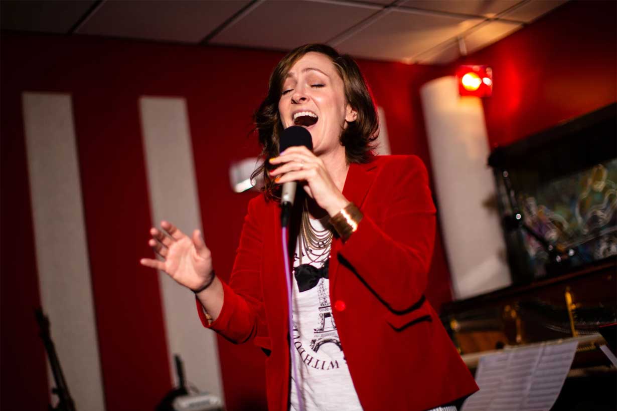 Sara Gazarek seen singing, eyes closed, holding a microphone, wearing a red jacket and white shirt