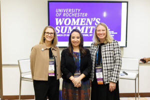 Three women stand side by side in front of a screen displaying “University of Rochester Women’s Summit,” posing after leading a session on board leadership at the 2025 event.