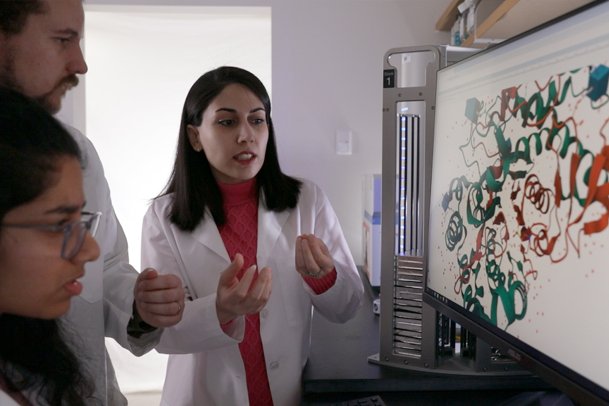 A woman in the white lab. coat looking at a monitor as a man and woman off to the side also look