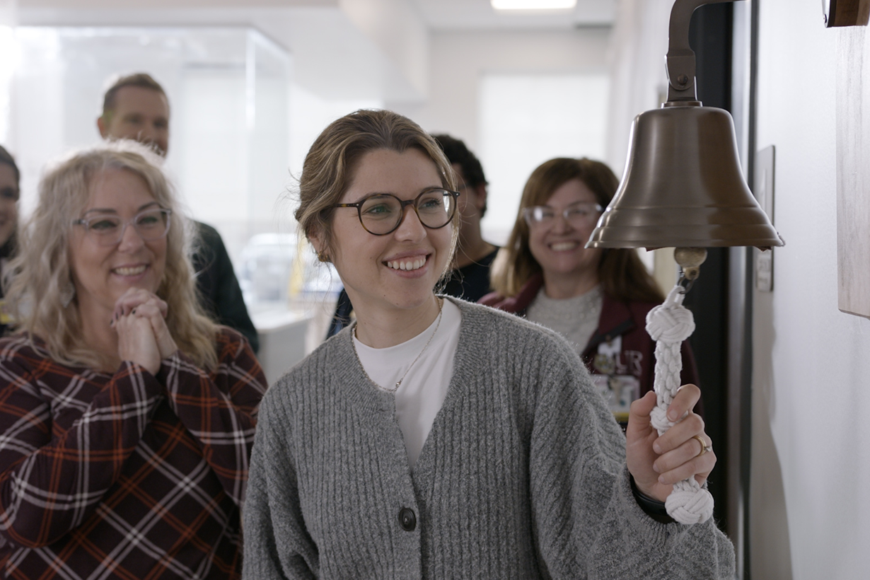 A girl with glasses smiling and looking off to the side. With people standing in the background
