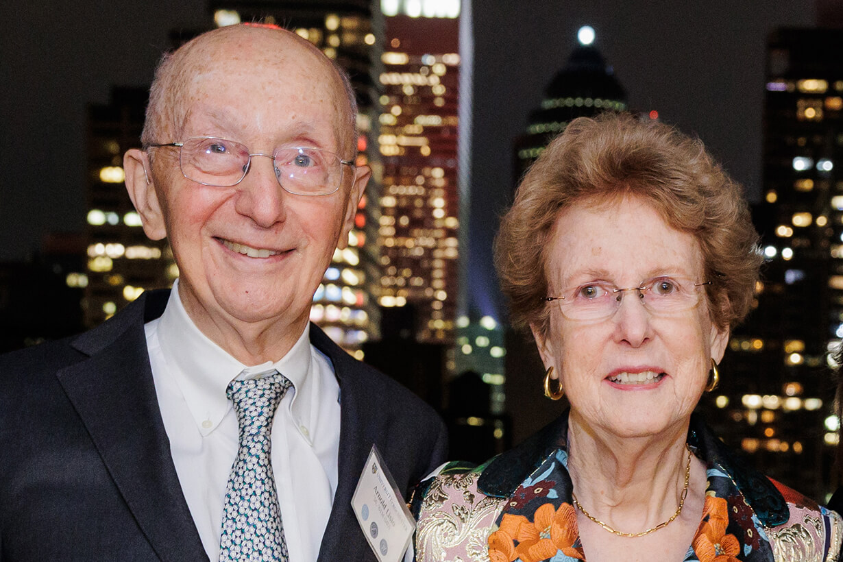 Arnold and Anne Lisio smiling at the camera, with a nighttime city skyline illuminated behind them.