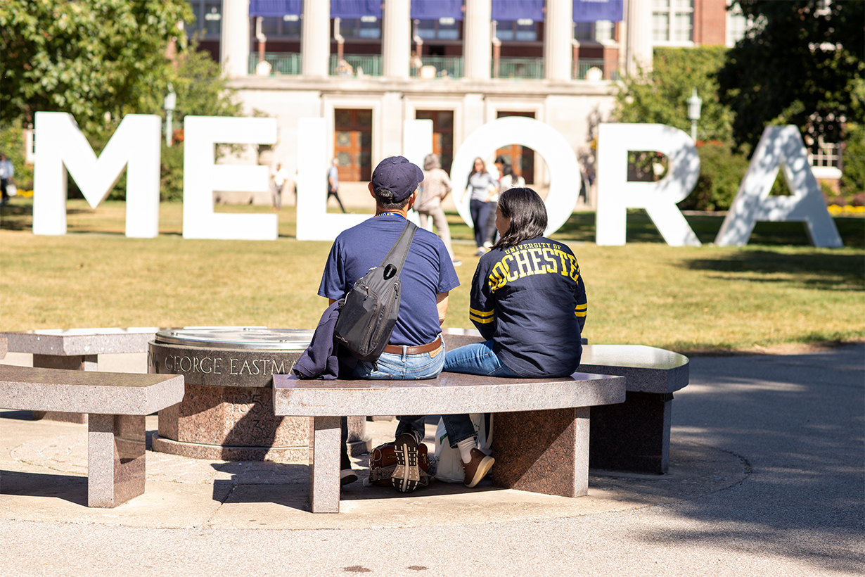 2 people sitting on a bench in the quad with their backs towards the camera and MELIORA letters in white in the background