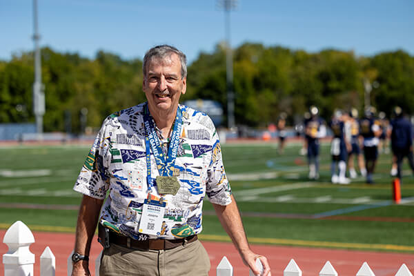 Joe Carson in a patterned shirt and khaki pants stands in front of a white picket fence at an athletic field, wearing lanyards and a medal from the National Senior Games.