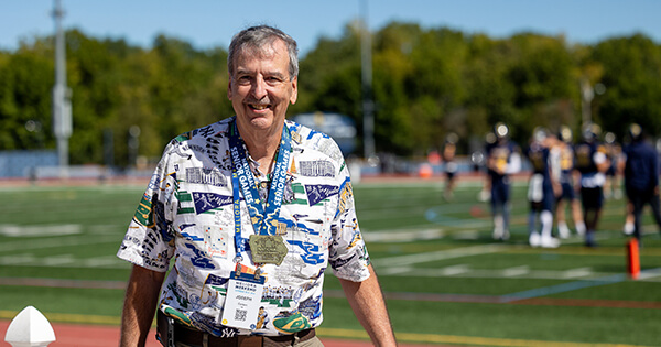 Joe Carson in a patterned shirt and khaki pants stands in front of a white picket fence at an athletic field, wearing lanyards and a medal from the National Senior Games.