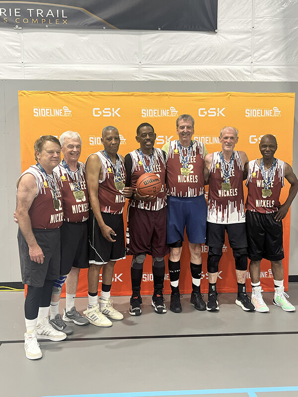 The Wazee Nickels senior basketball team poses together in their maroon and white jerseys, proudly displaying gold medals around their necks following a competition.