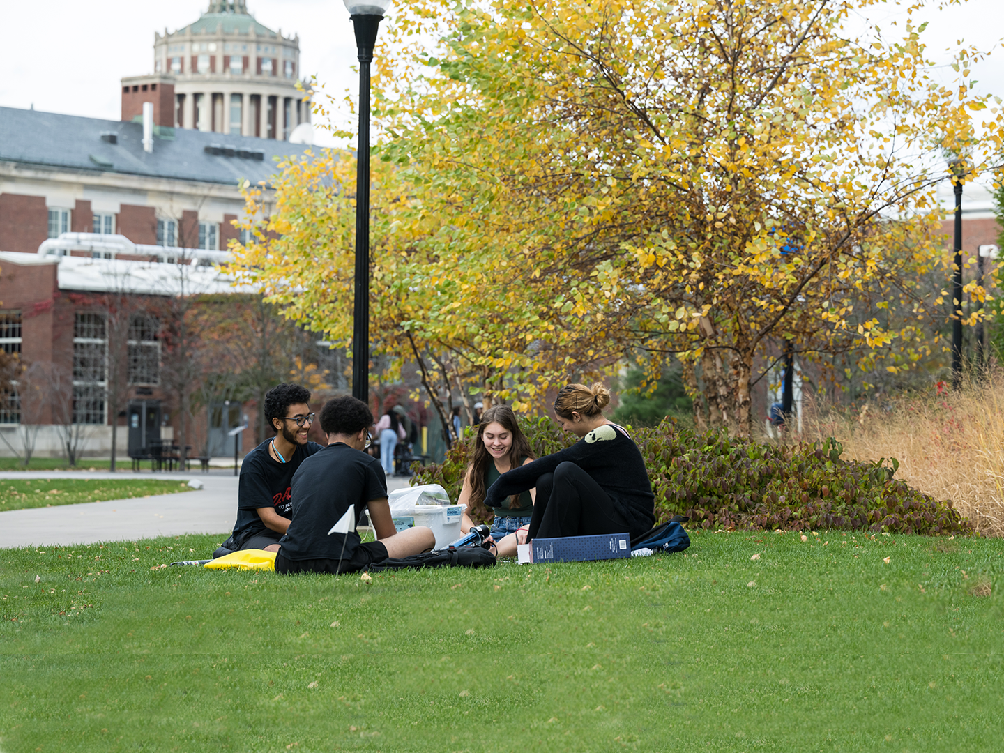 Group of 4 students sitting on the lawn with Rush Rhees library tower in background