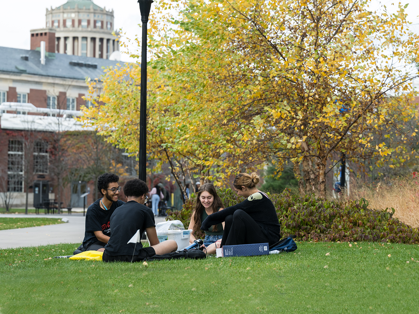 Group of 4 students sitting on the lawn with Rush Rhees library tower in background