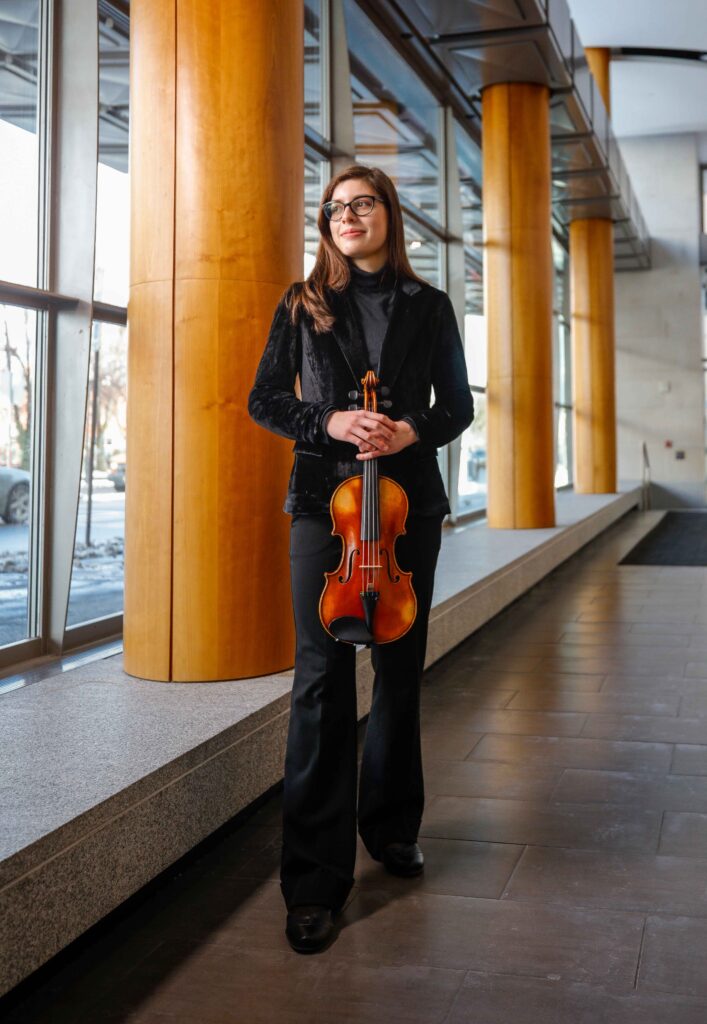 Sofia Grimes '27E hodling her violin in a hallway with windows on left side of photo