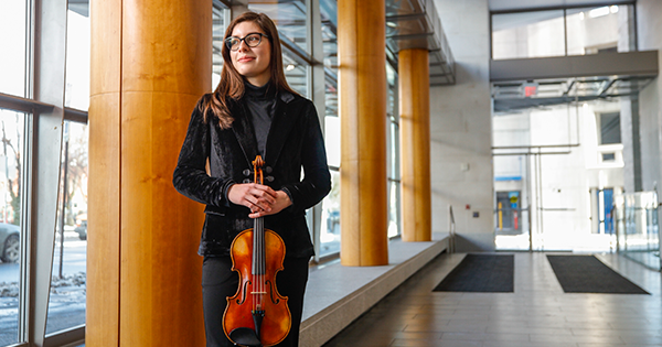 Sofia Grimes '27E holding her violin in a hallway with windows on left side of photo
