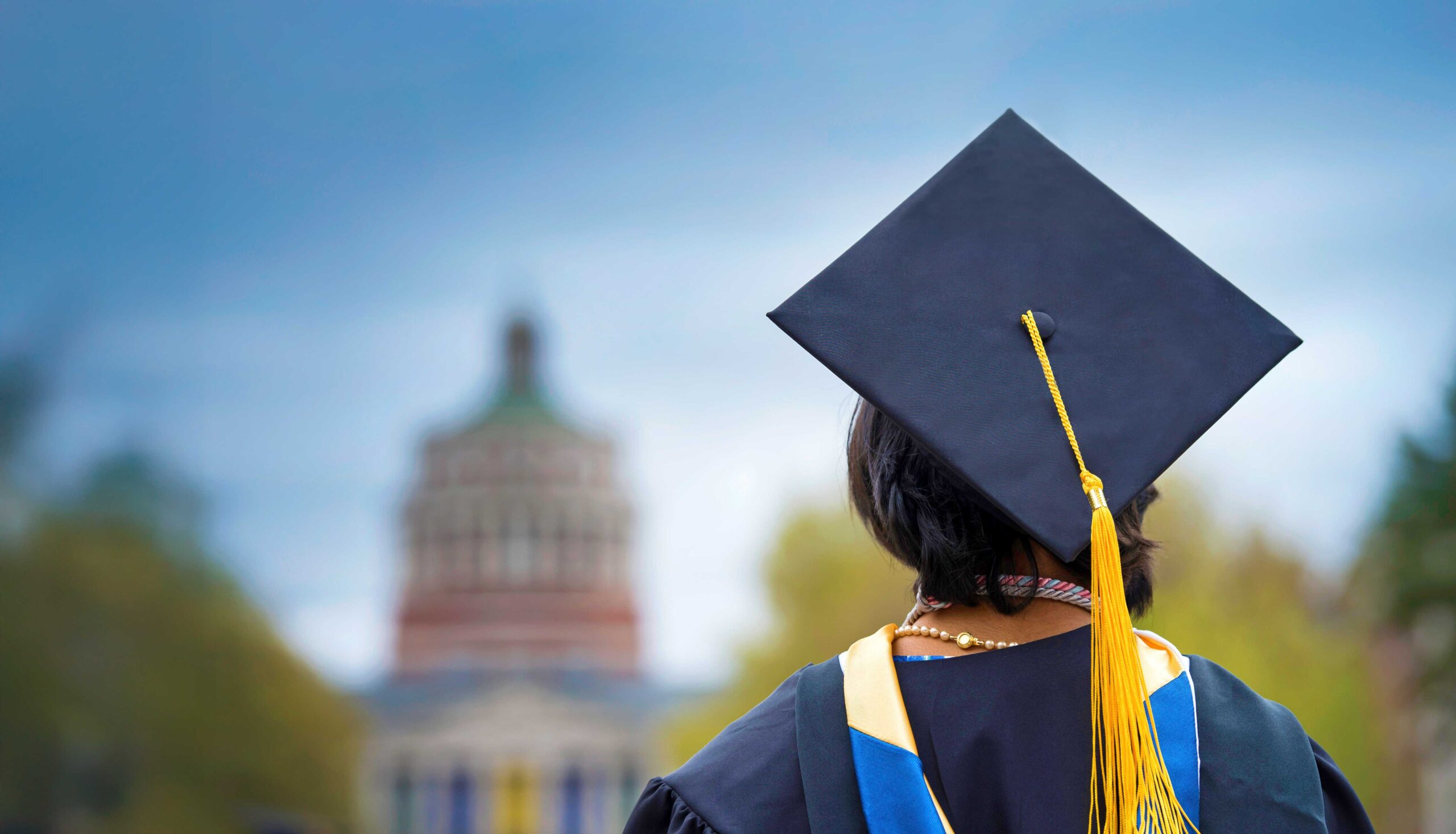 The back of a student in cap and gown in the foreground looking towards Rush Rhees library tower in the background blurred out against a blue sky