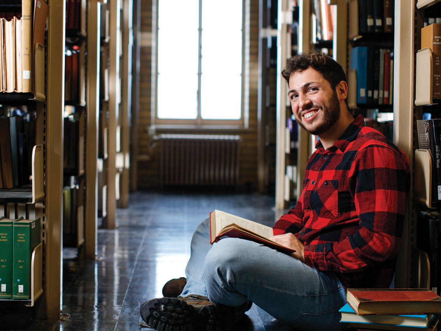 Jacob Souza sitting on the ground between library stack with a book in lap smiling at the camera