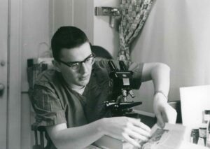 A young Alexander “Al” A. Levitan black and white photo wearing glasses with a microscope in front of him examining a piece a document